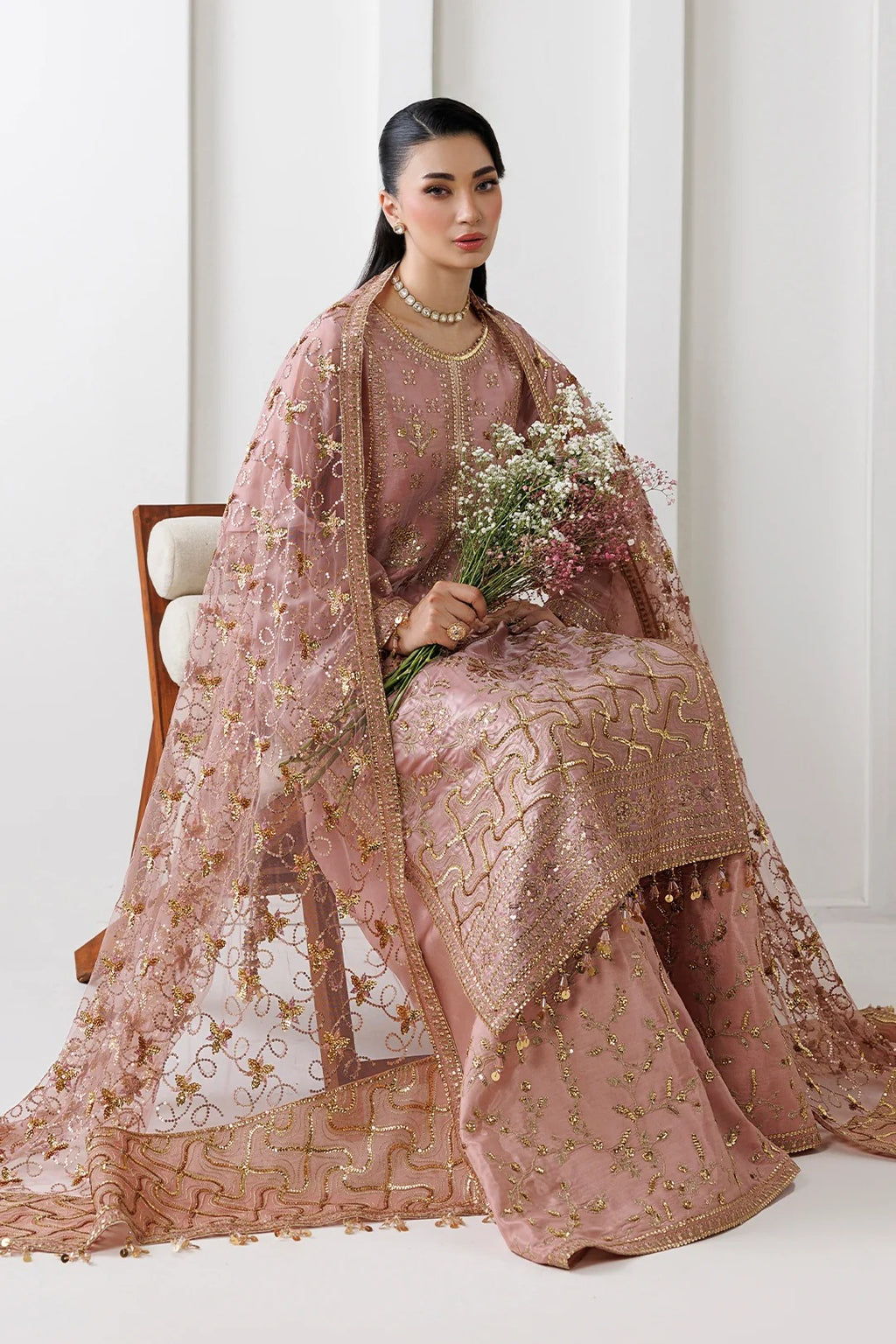 A woman seated on a wooden chair wearing an ornate dusty pink traditional outfit with gold embroidery and a matching sheer embroidered dupatta, holding a bouquet of flowers in a bright indoor setting.