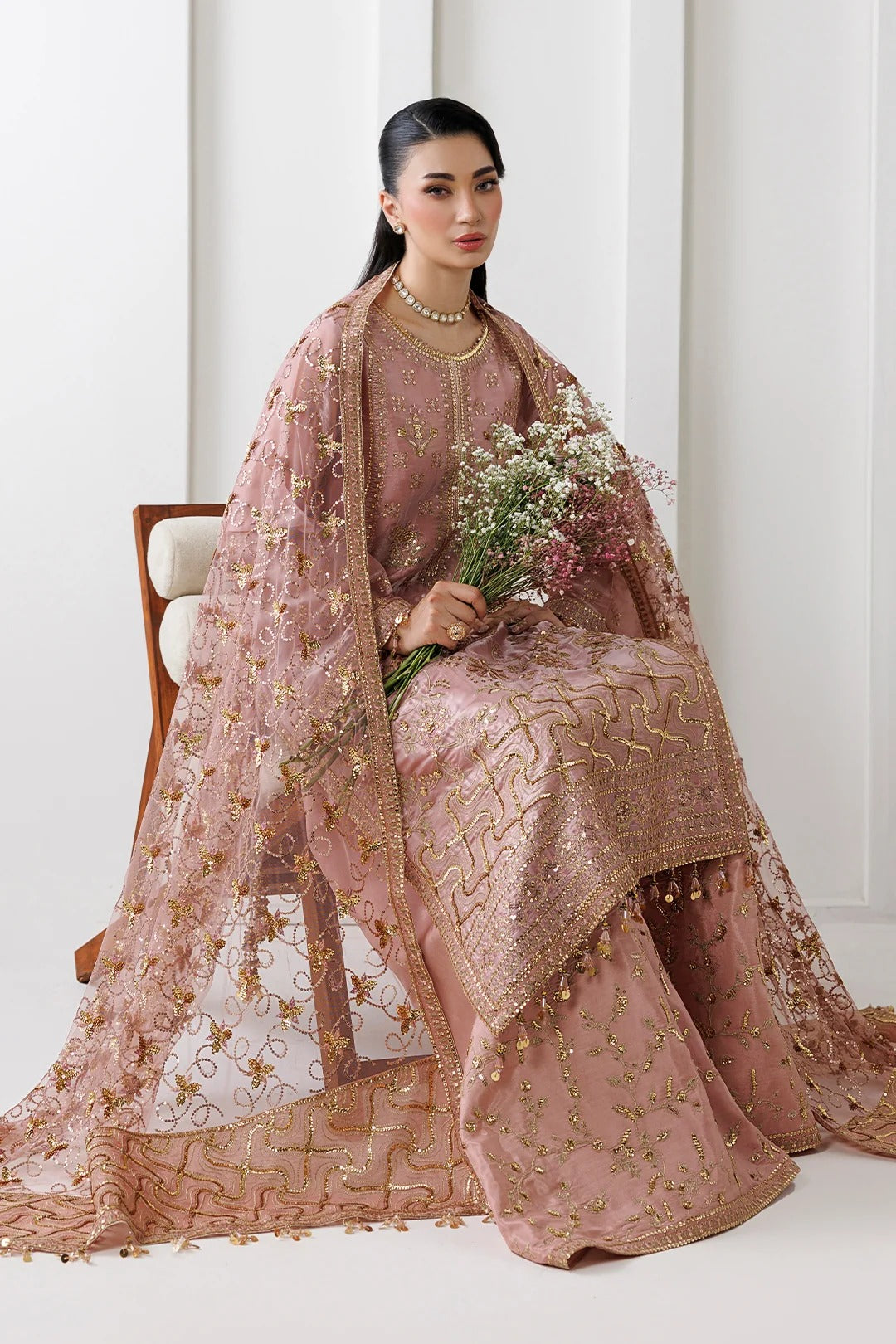 A woman seated on a wooden chair wearing an ornate dusty pink traditional outfit with gold embroidery and a matching sheer embroidered dupatta, holding a bouquet of flowers in a bright indoor setting.