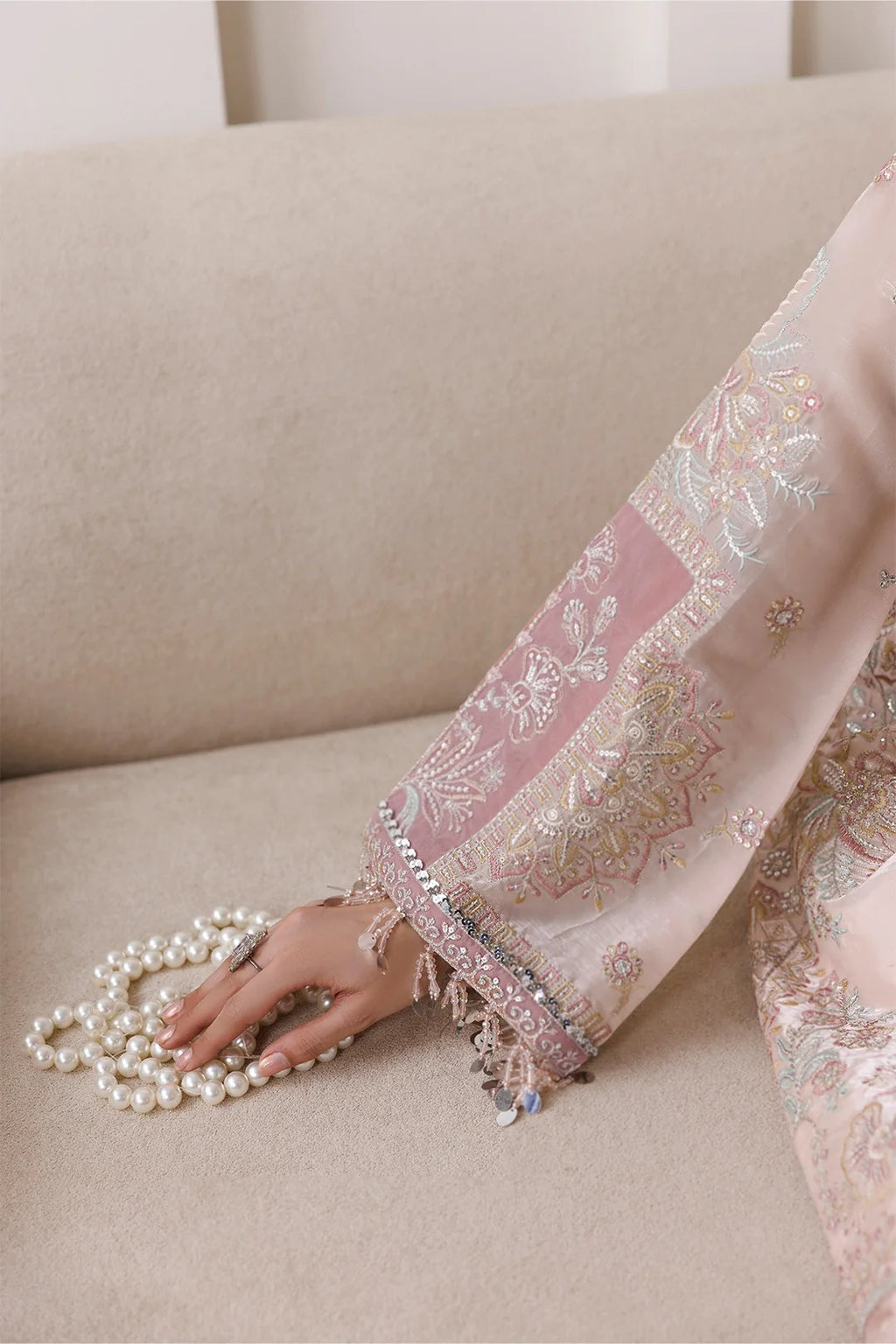 A close-up of a hand resting on a beige sofa next to a strand of white pearls, wearing a light pink embroidered traditional garment with metallic gold and silver floral patterns.