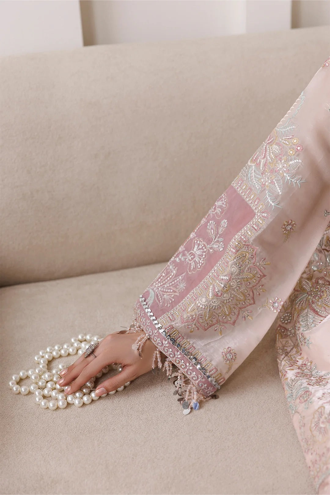 A close-up of a hand resting on a beige sofa next to a strand of white pearls, wearing a light pink embroidered traditional garment with metallic gold and silver floral patterns.