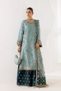 A woman models a traditional three-piece ethnic outfit consisting of a light blue embroidered kurta, a matching dupatta, and dark teal wide-leg pants with gold floral motifs. She is holding a matching light blue embroidered clutch bag against a clean white studio background.