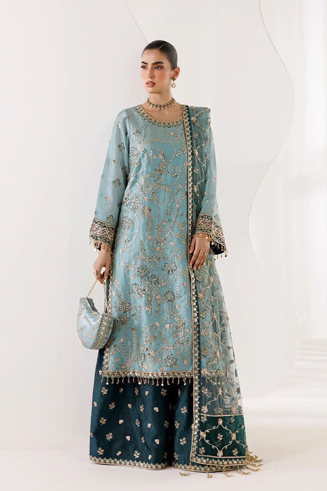 A woman models a traditional three-piece ethnic outfit consisting of a light blue embroidered kurta, a matching dupatta, and dark teal wide-leg pants with gold floral motifs. She is holding a matching light blue embroidered clutch bag against a clean white studio background.