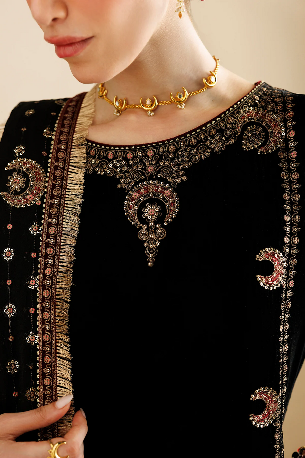 Close-up of a model wearing a black embroidered traditional outfit with ornate gold and pink embellishments, a gold necklace, and a gold ring against a soft beige background.