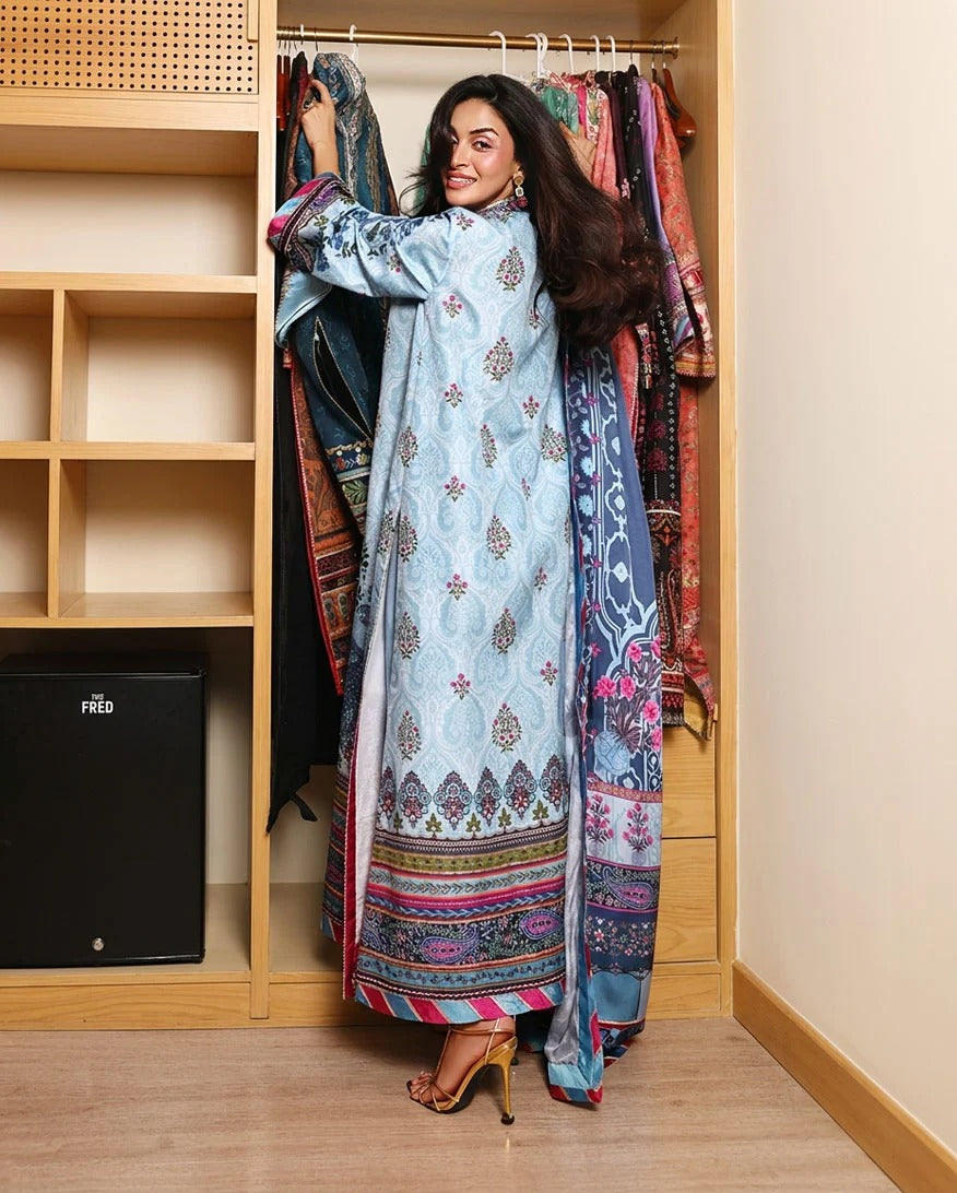 A woman stands in front of an open wooden closet, holding up a light blue embroidered traditional outfit with a matching patterned dupatta. The outfit features intricate gold, pink, and green embroidery, and is styled with gold high-heeled sandals.