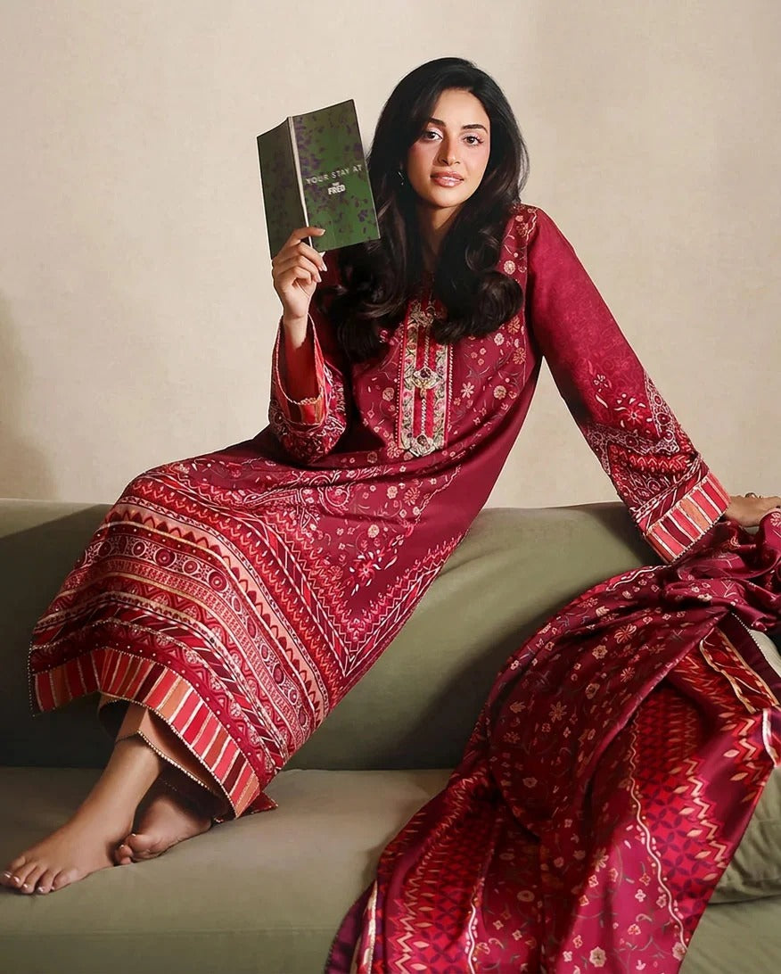 A woman seated on a sofa wearing a deep red embroidered traditional outfit with matching draped fabric, holding a book against a neutral beige wall.