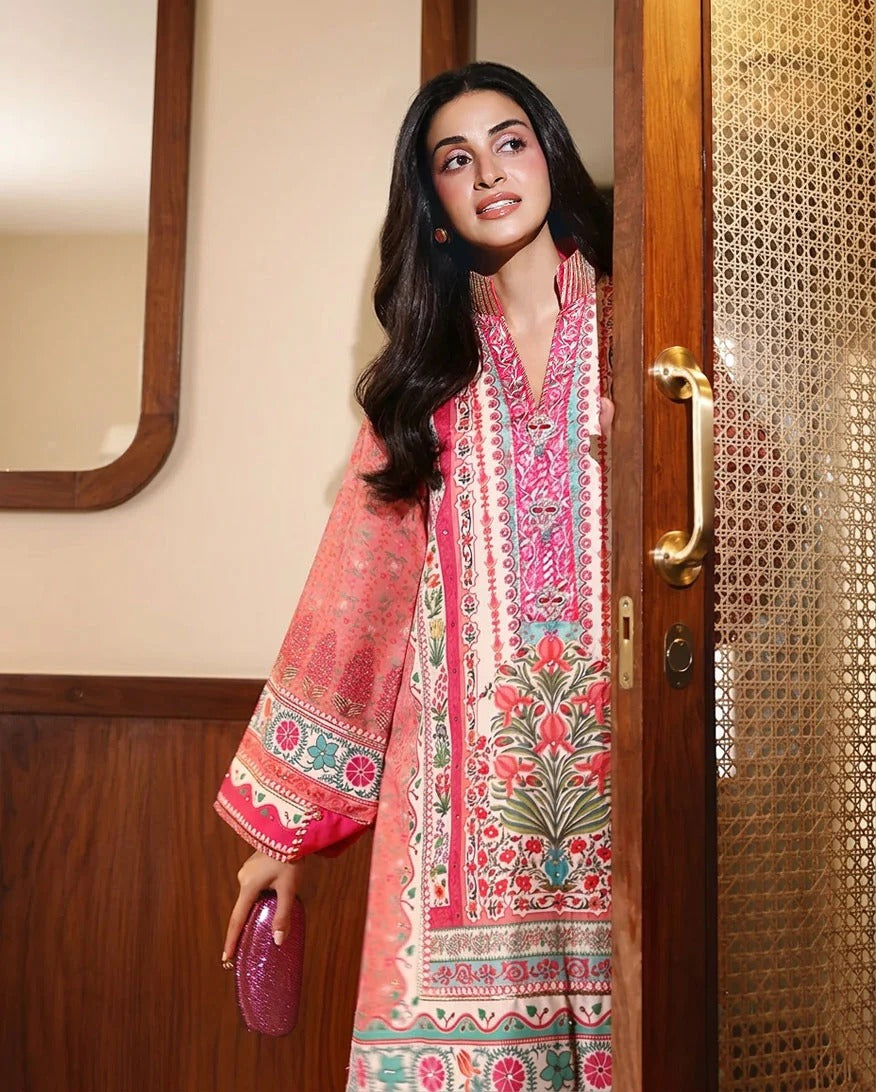 A woman wearing a vibrant pink and multicolor printed traditional South Asian kurta stands in a doorway holding a matching pink clutch bag.