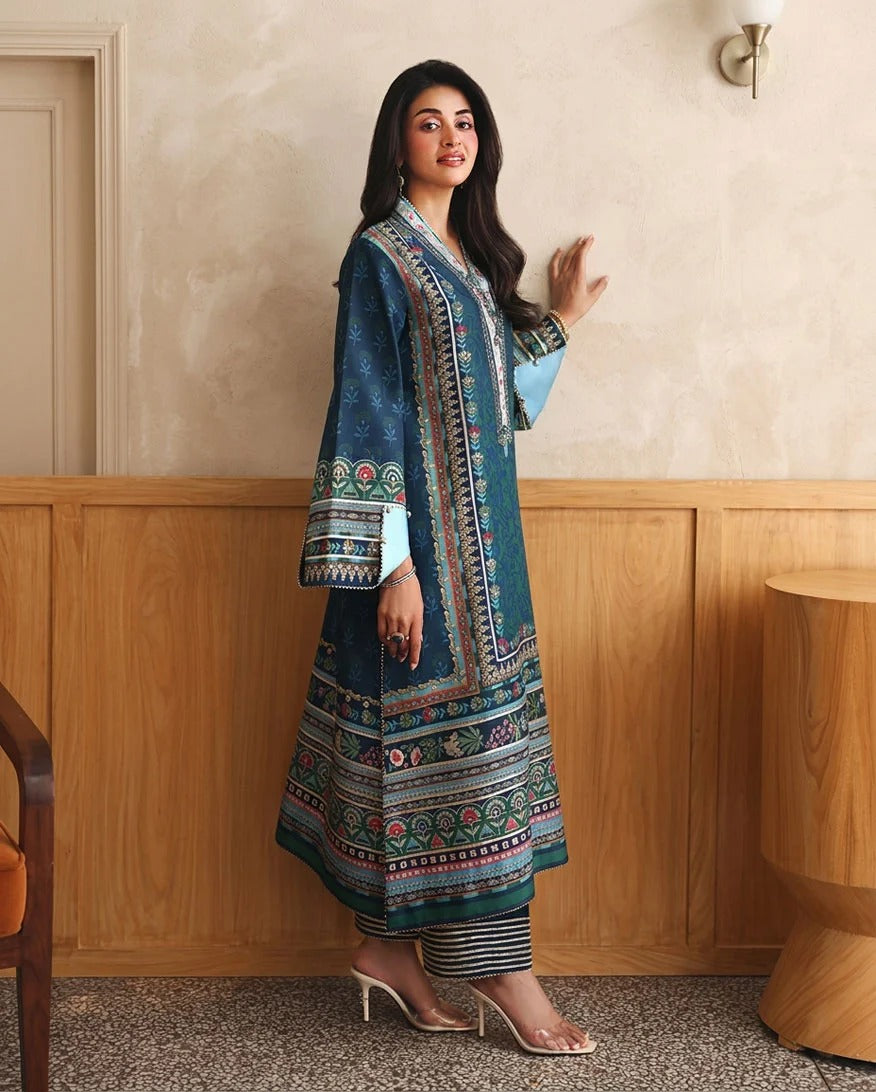A woman models a traditional South Asian outfit featuring a teal blue printed kurta with intricate multicolor patterns and a light blue inner kurta, paired with striped trousers and clear high-heeled sandals, set against a warm beige wall and wooden paneling.