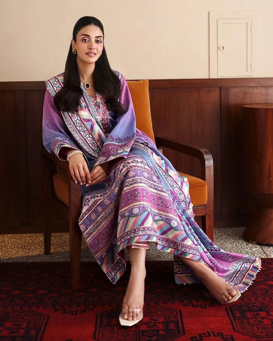 A woman sits in an orange armchair wearing a purple and multicolor patterned traditional outfit with a matching draped shawl. The setting features a dark wood-paneled wall, a red patterned rug, and a cream-colored wall in the background.