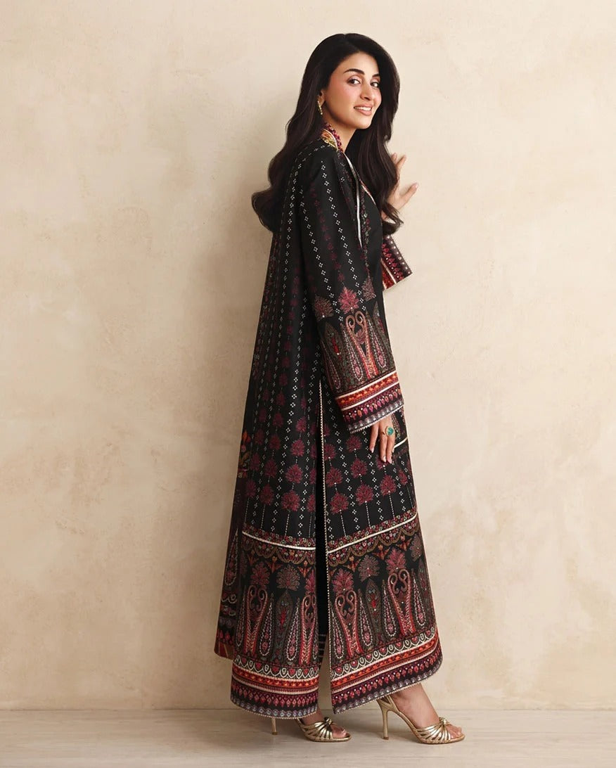 A woman modeling a long black printed kurta with red and beige patterns, standing against a textured beige wall.