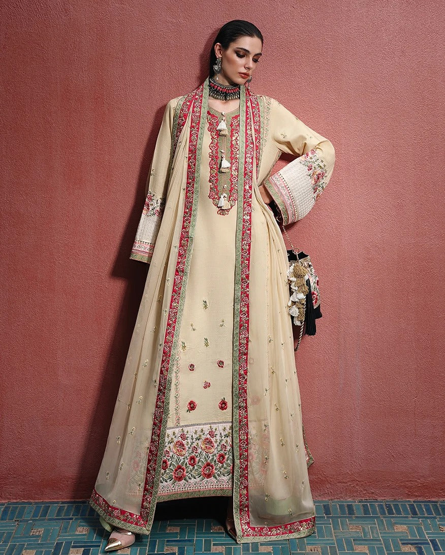 A woman models a traditional beige Anarkali suit with red and green floral embroidery, a matching dupatta, and a long beige coat with red borders, standing against a textured reddish-brown wall.