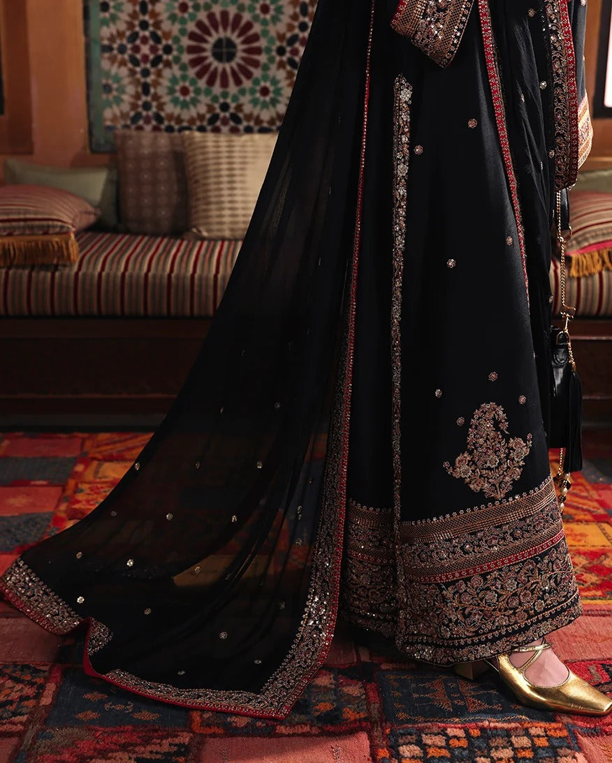 A close-up of a person wearing a black traditional embroidered outfit with a matching sheer dupatta, standing on a patterned rug in an indoor setting with a sofa and wall hanging visible in the background.