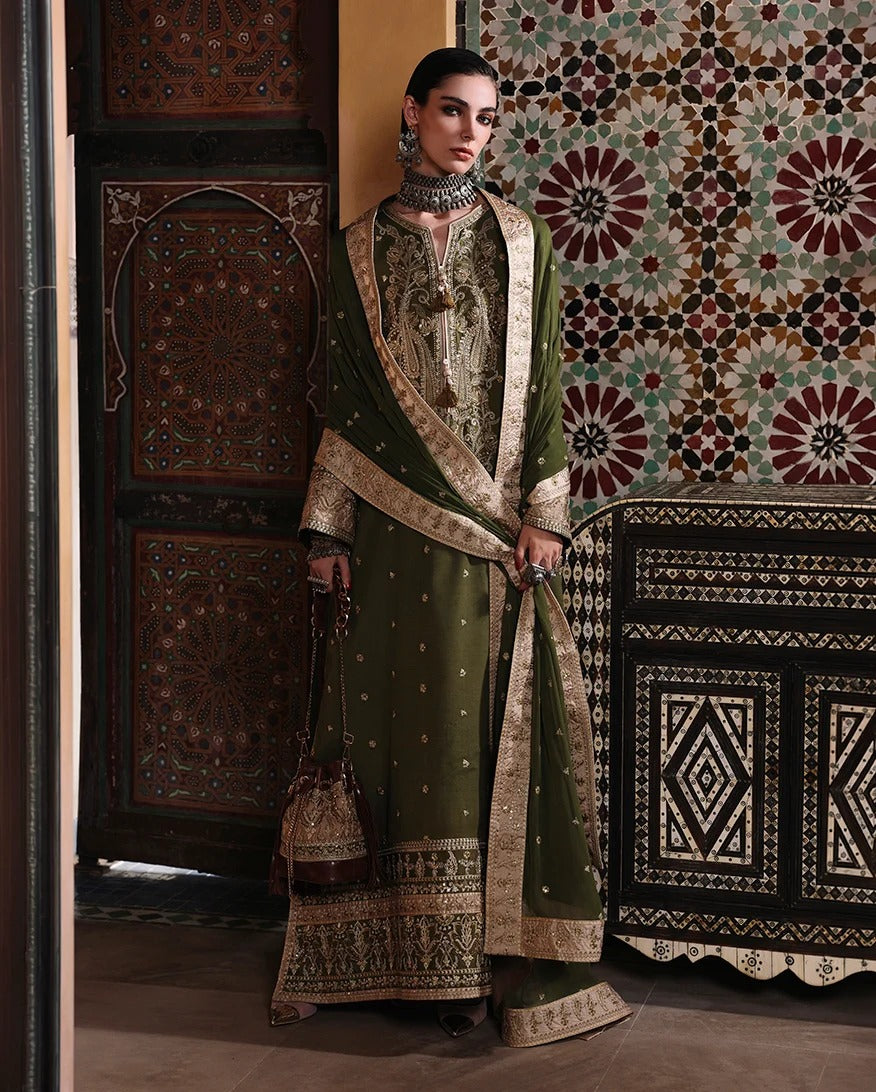 A woman models an olive green traditional South Asian outfit with intricate gold embroidery, a matching dupatta, and a small brown handbag, set against a background of ornate wooden doors and a patterned tapestry.