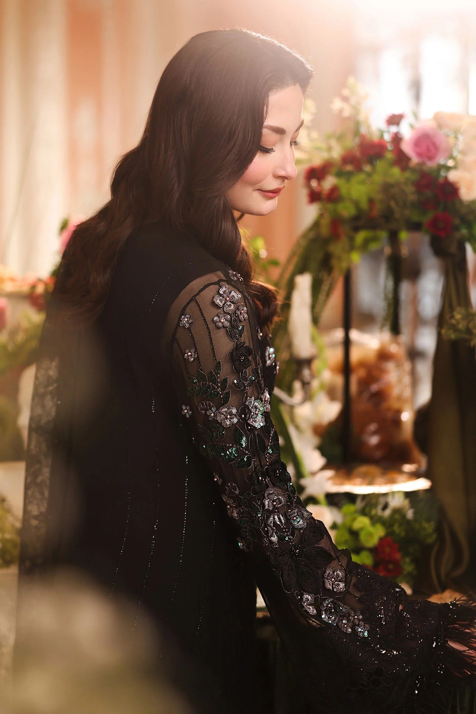 A woman in a black embellished long-sleeve dress with sheer lace sleeves, photographed in a warm, softly lit indoor setting with blurred flowers in the background.