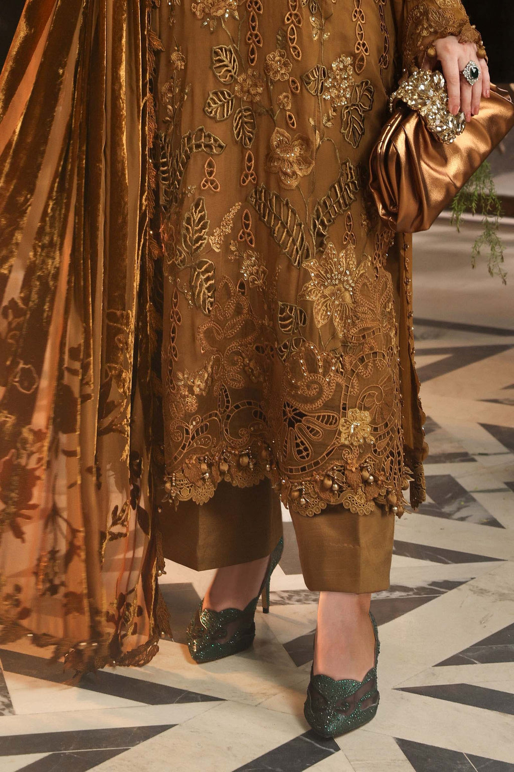 A close-up of a person wearing a brown embroidered traditional outfit with a matching dupatta, holding a metallic clutch, and wearing dark green embellished heels on a patterned floor.