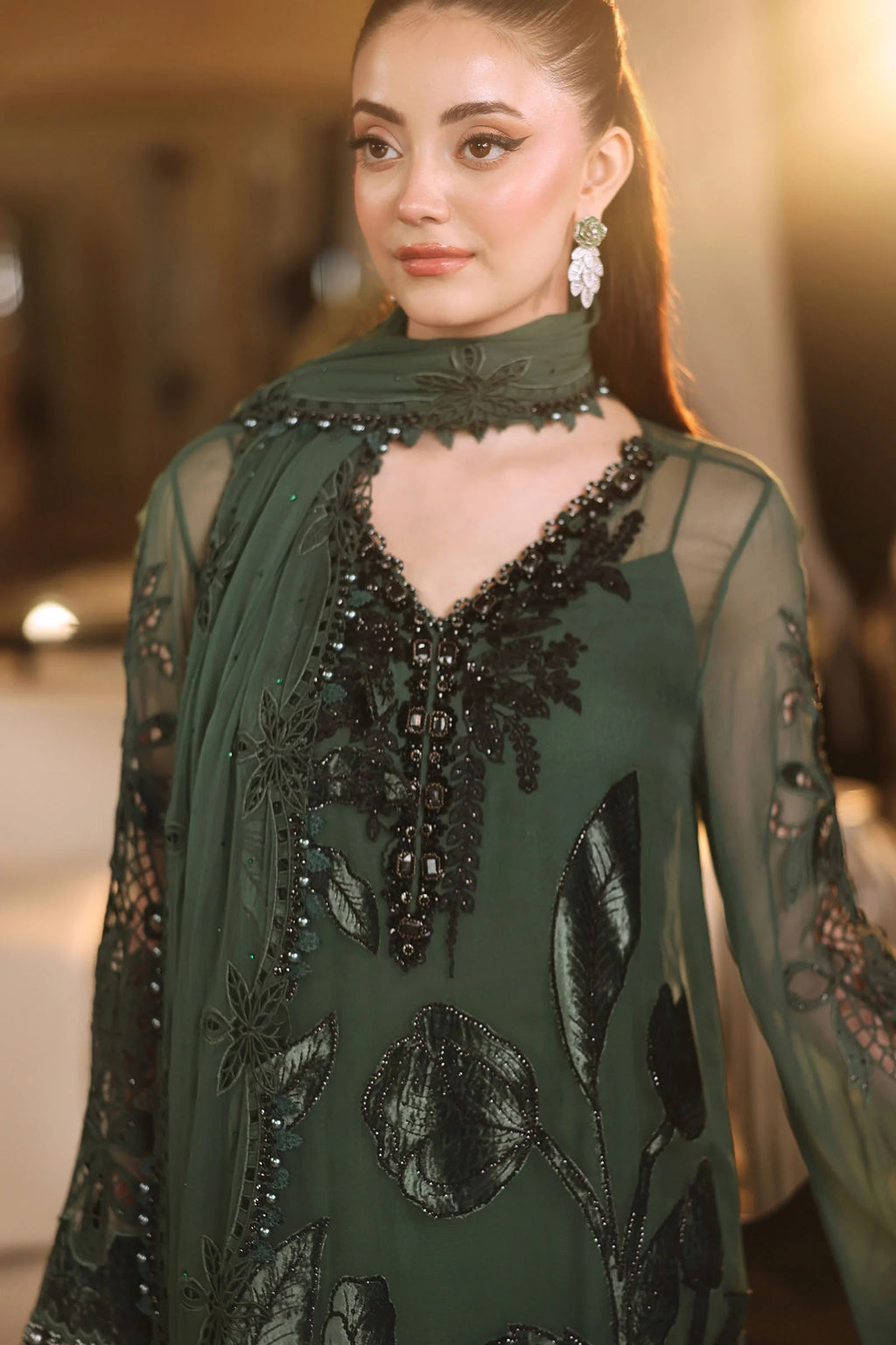 A woman wearing a dark green embroidered traditional outfit with sheer sleeves, black floral embroidery, and a matching dupatta, photographed in a softly lit indoor setting.