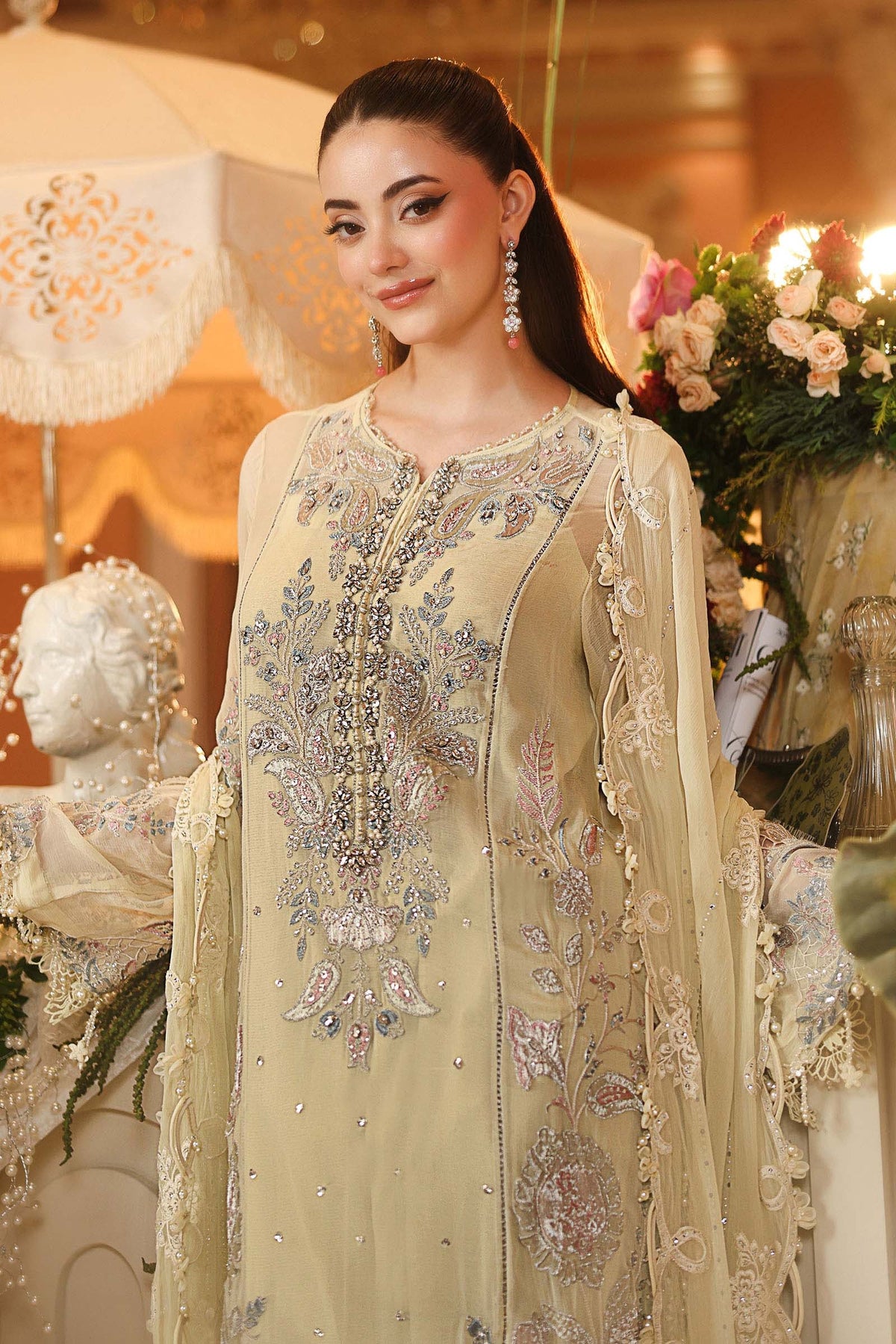 A woman wearing an ornate beige traditional outfit with intricate silver and blue embroidery, standing in a decorated indoor setting with flowers and draped fabric in the background.