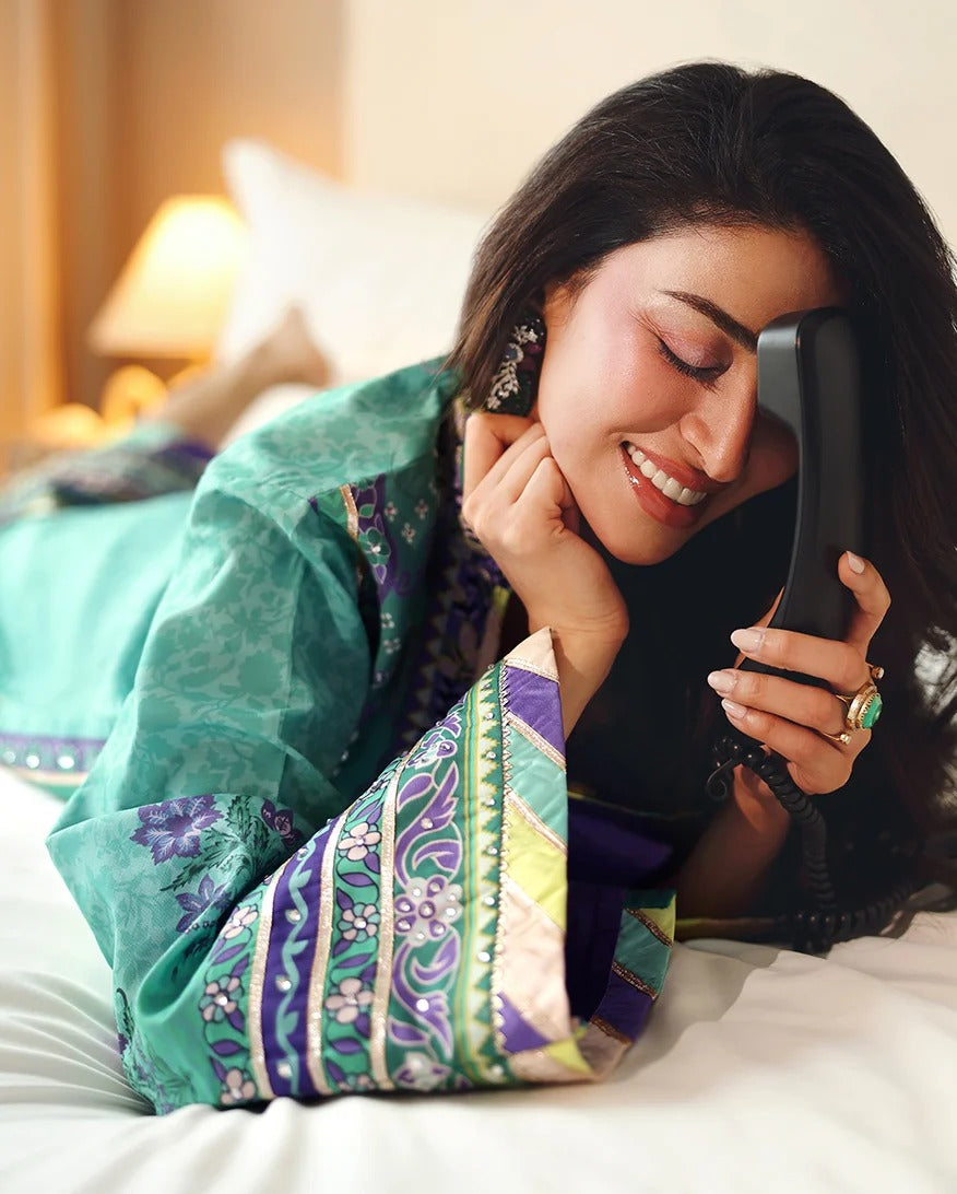 A woman lying on a bed in a teal and purple patterned kurta, smiling while holding a black cordless phone receiver.