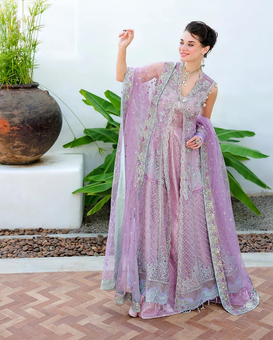 A woman models a lavender purple traditional Indian outfit with intricate silver embroidery, featuring a heavily embellished dupatta draped over her shoulders, set against a white wall with potted plants.