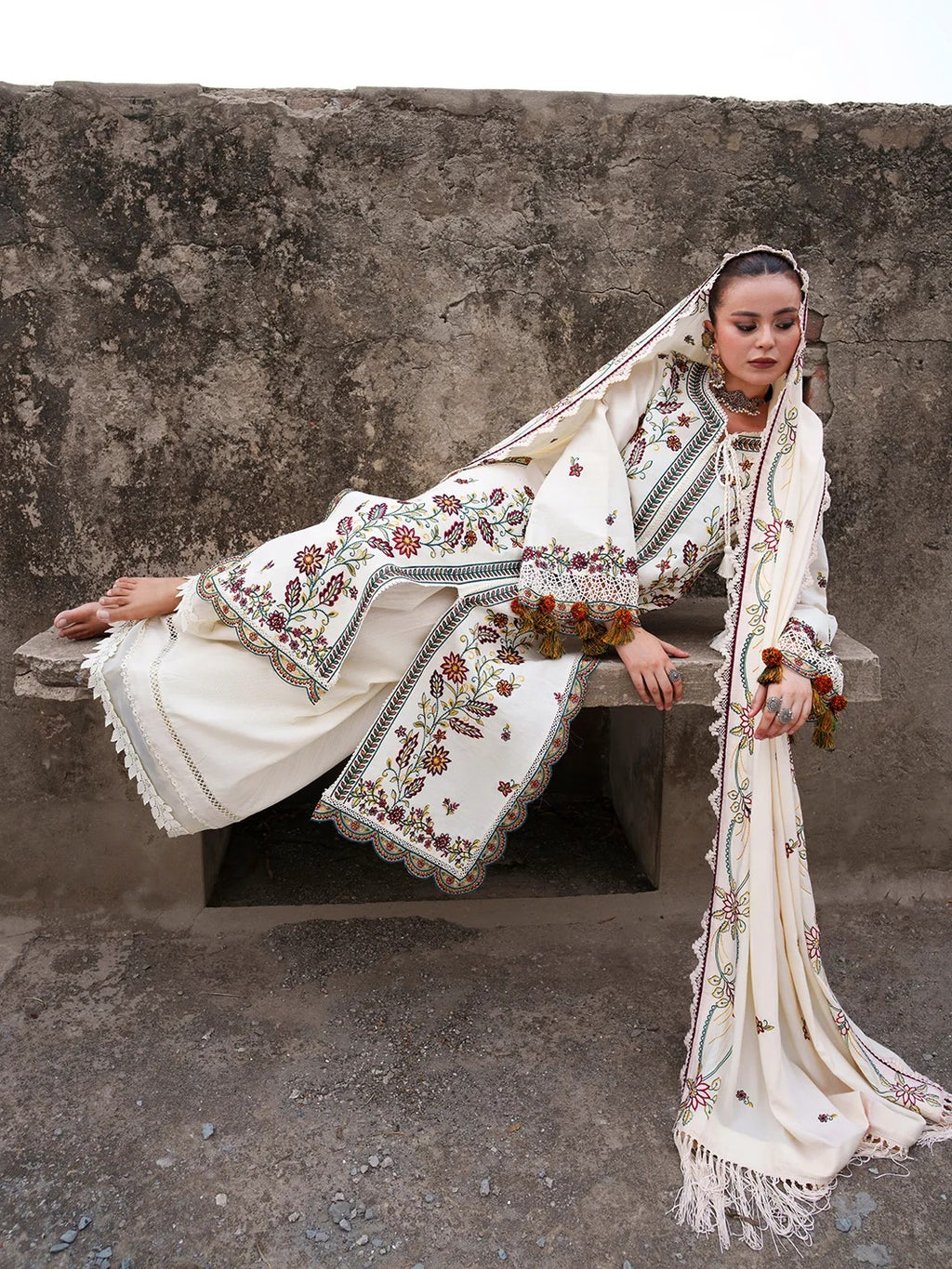 A woman reclines on a stone bench against a textured concrete wall, wearing an ornate cream-colored traditional outfit with intricate multicolor floral embroidery and a matching embroidered dupatta.