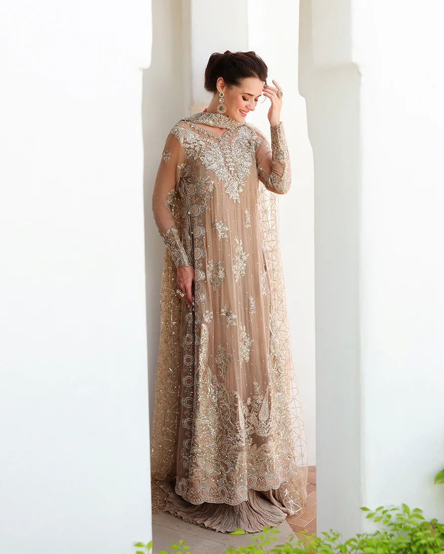 A woman wearing a long beige embellished gown with intricate silver and gold embroidery, standing in a bright white architectural setting.