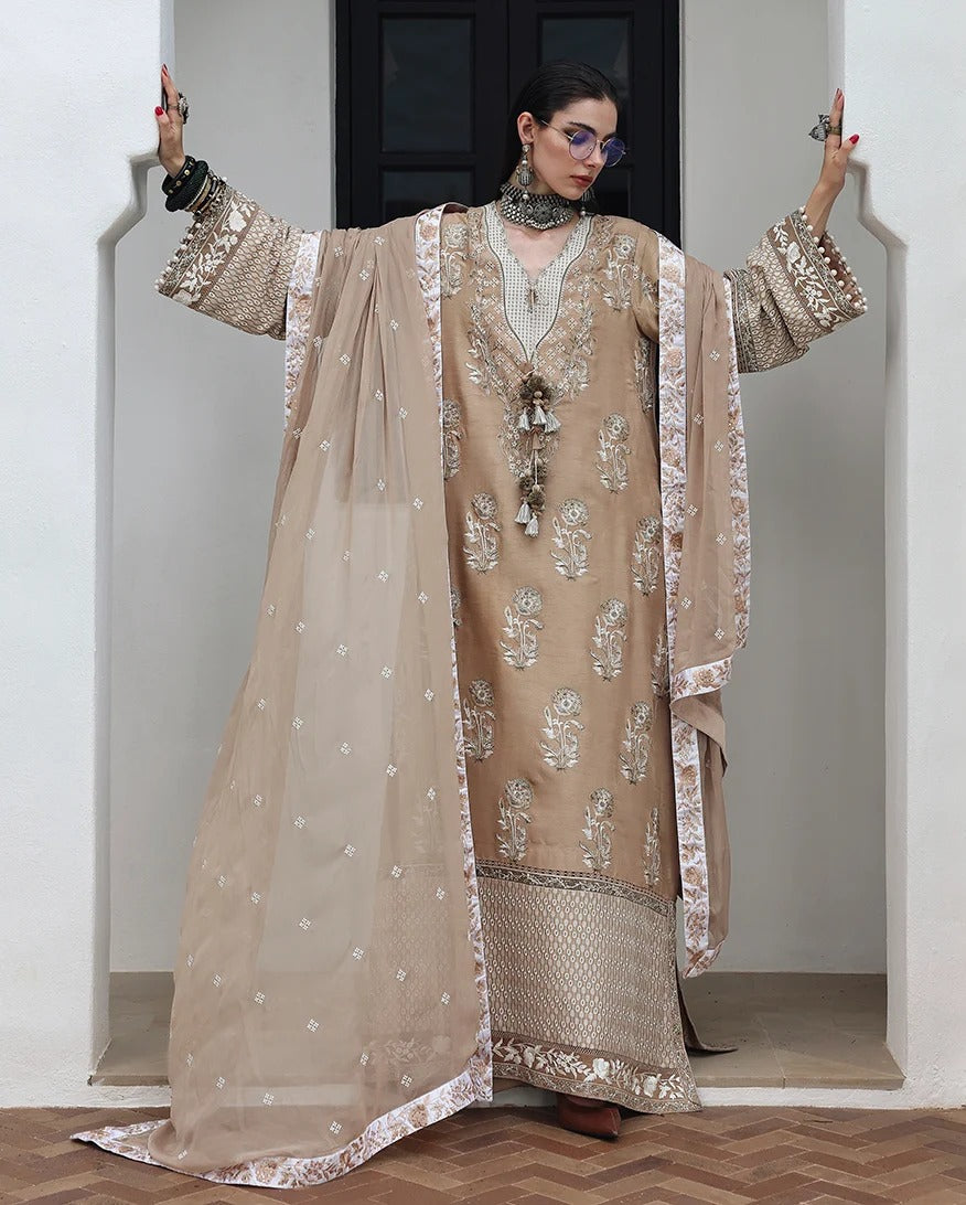 A model wearing a beige and gold embroidered traditional South Asian outfit with a matching sheer dupatta, standing in a doorway with arms raised.