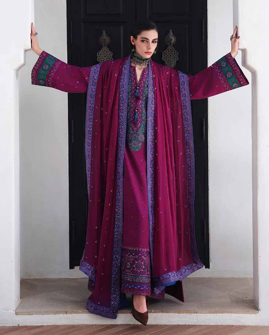 A woman models a traditional deep magenta three-piece outfit with ornate blue and green embroidery, featuring a matching draped outer layer, standing in front of a black door.