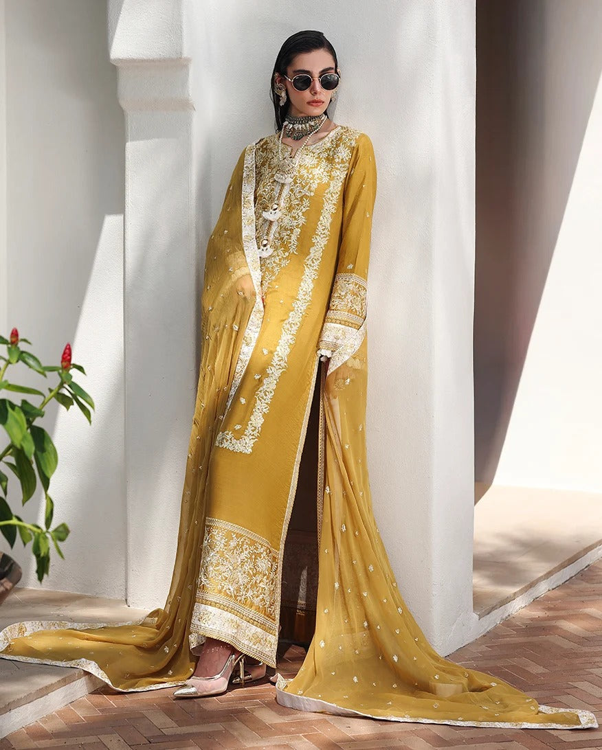 A woman models a traditional South Asian salwar kameez suit in mustard yellow with intricate white and gold embroidery, paired with a matching sheer dupatta, set against a white architectural background.