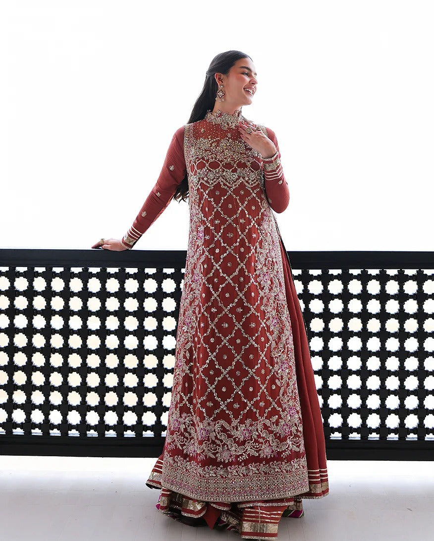 A woman models a traditional Indian salwar kameez outfit with a rust-red base and intricate silver mirror work embroidery, standing against a white background with a black decorative railing.
