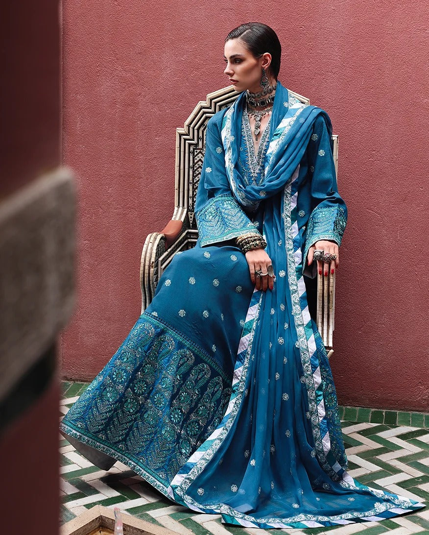 A woman seated on a decorative chair wearing a rich blue embroidered traditional outfit with a matching draped scarf, set against a textured reddish-brown wall and patterned tile floor.