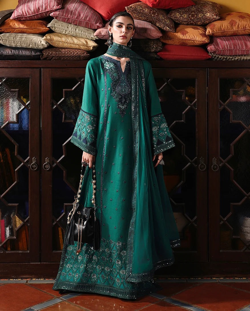 A woman models a traditional green embroidered dress with a matching sheer dupatta and black handbag, standing in front of a dark wooden cabinet with stacked cushions.