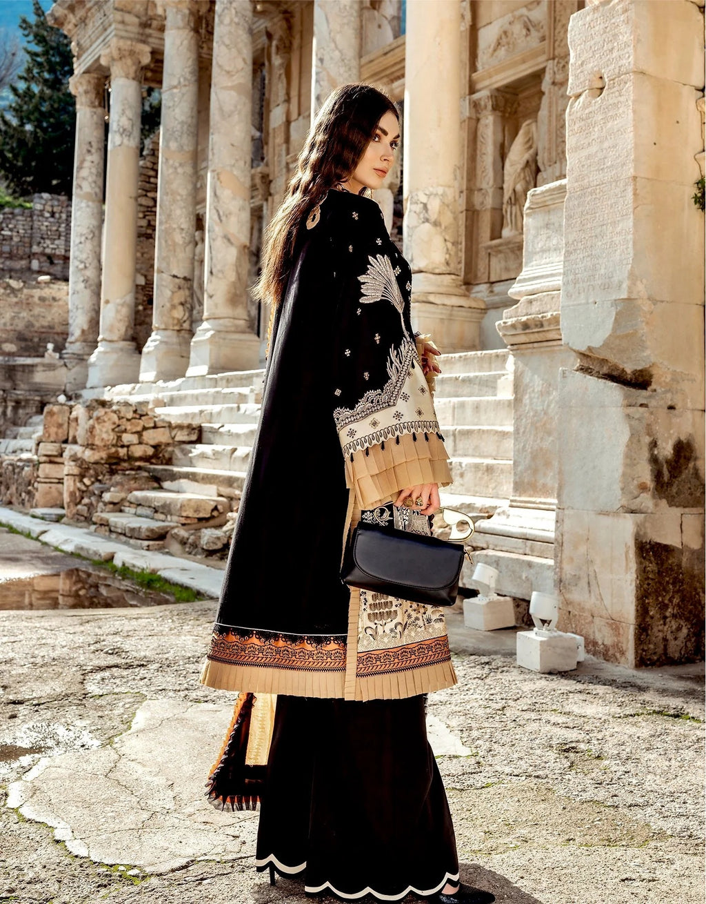 A woman stands in front of ancient stone columns and ruins, wearing a black traditional outfit with beige embroidered sleeves and a decorative beige border, holding a black handbag.