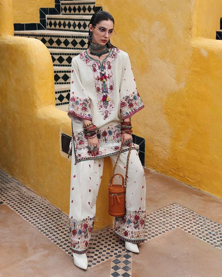 A model wearing a cream-colored embroidered traditional outfit with wide-leg pants, a matching embroidered kurta, and white pointed shoes, holding a brown leather bucket bag, set against a yellow stucco wall and patterned tile stairs.