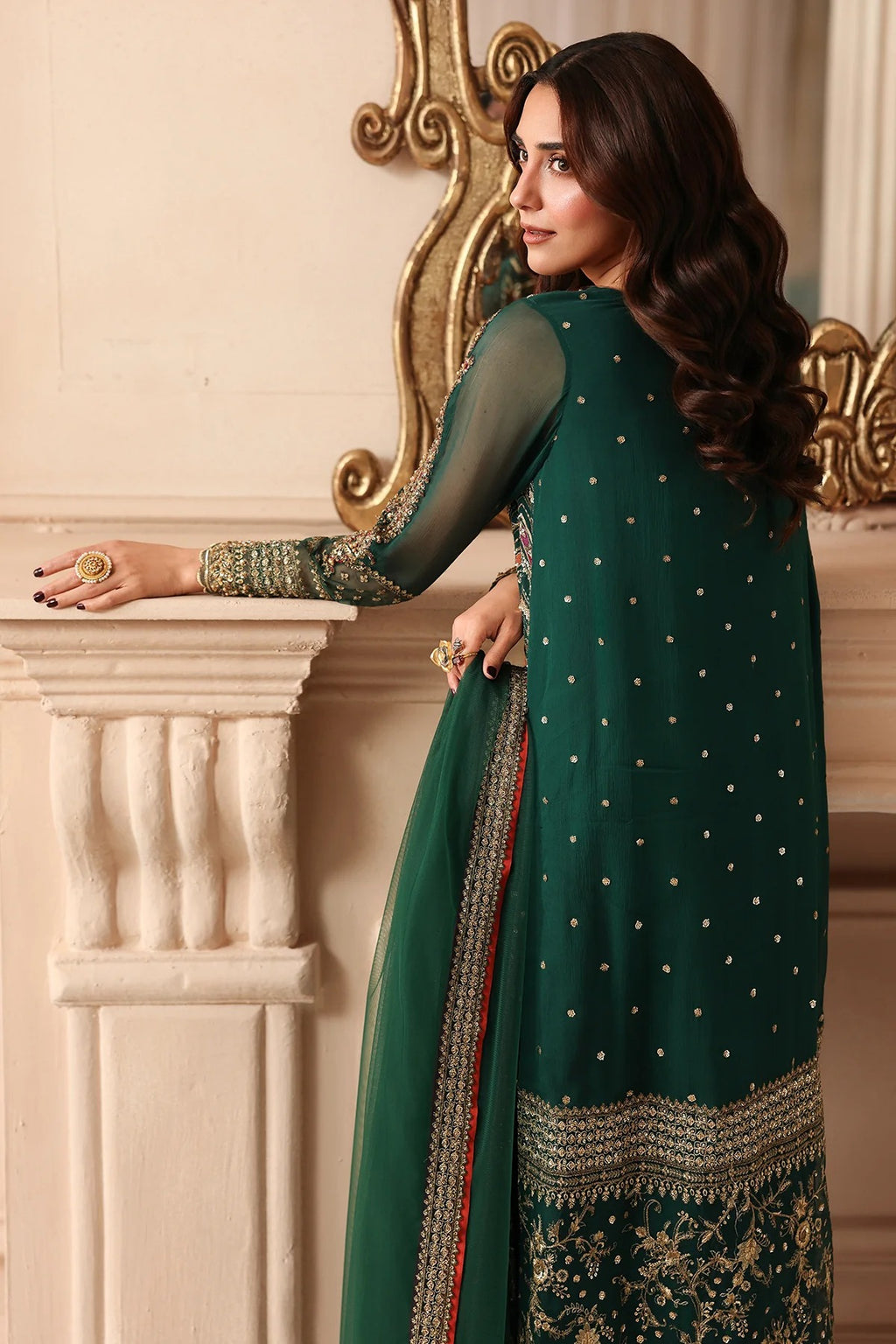 A woman wearing a dark green traditional outfit with gold embroidery and sheer sleeves, standing indoors near a white column and ornate mirror.