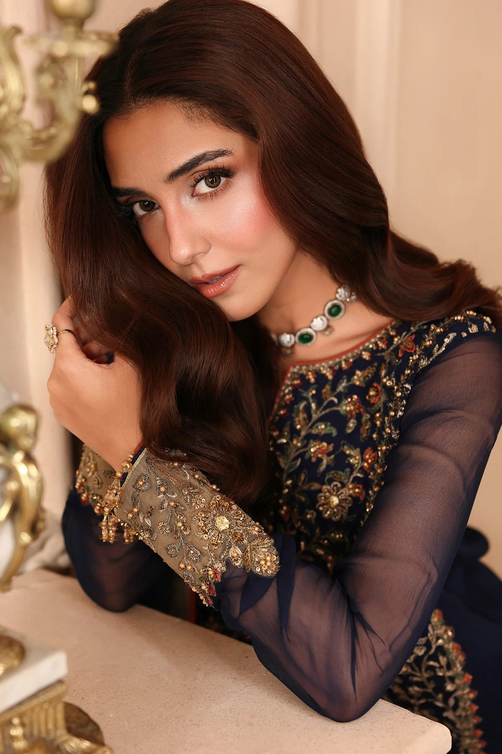 A woman with long dark brown hair models an elegant navy blue traditional outfit with intricate gold embroidery and sheer sleeves, accessorized with a green and white beaded necklace.