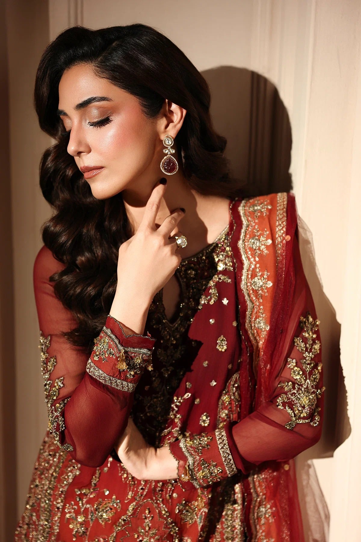 A woman wearing a rich red and gold embroidered traditional outfit with sheer sleeves and ornate jewelry, posed against a warm beige wall.