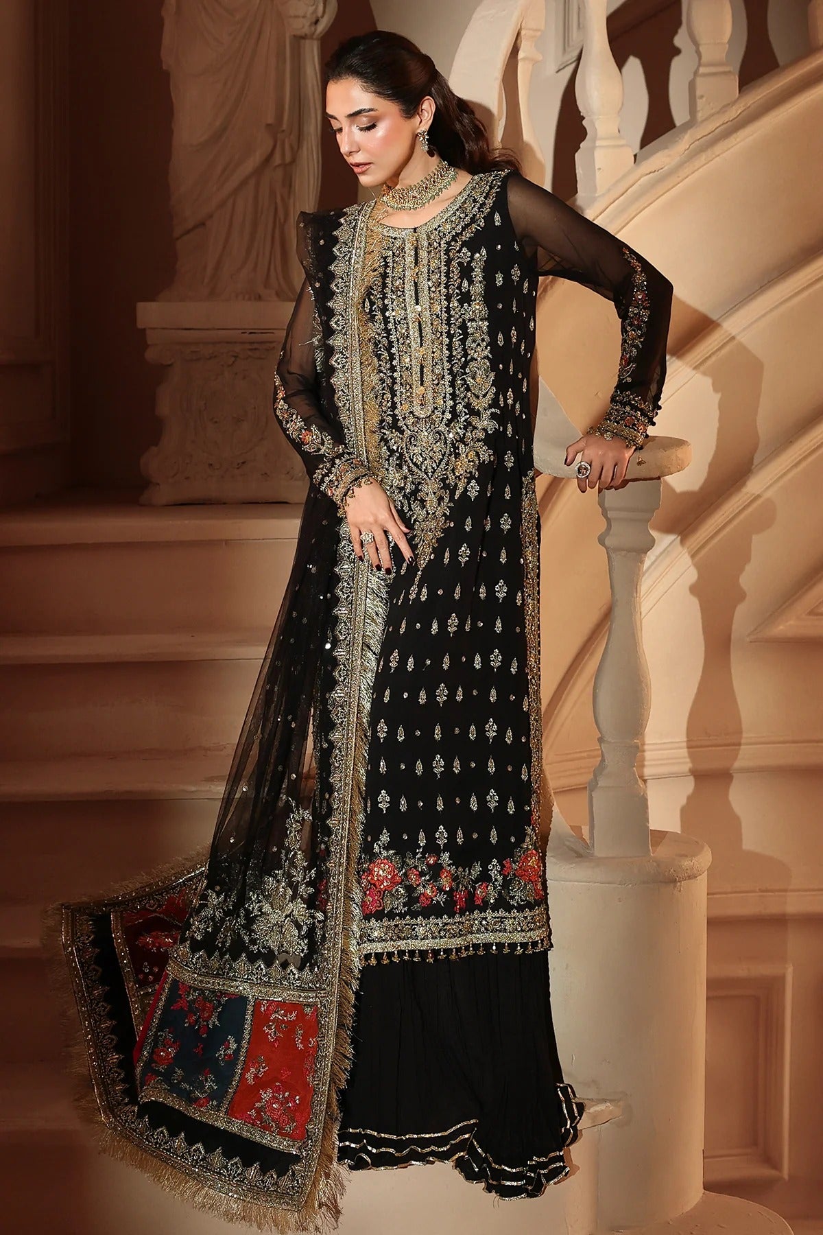 A woman models a black traditional embroidered outfit with sheer sleeves, heavy gold and silver embroidery, and a matching patterned dupatta in an elegant indoor setting with a staircase.