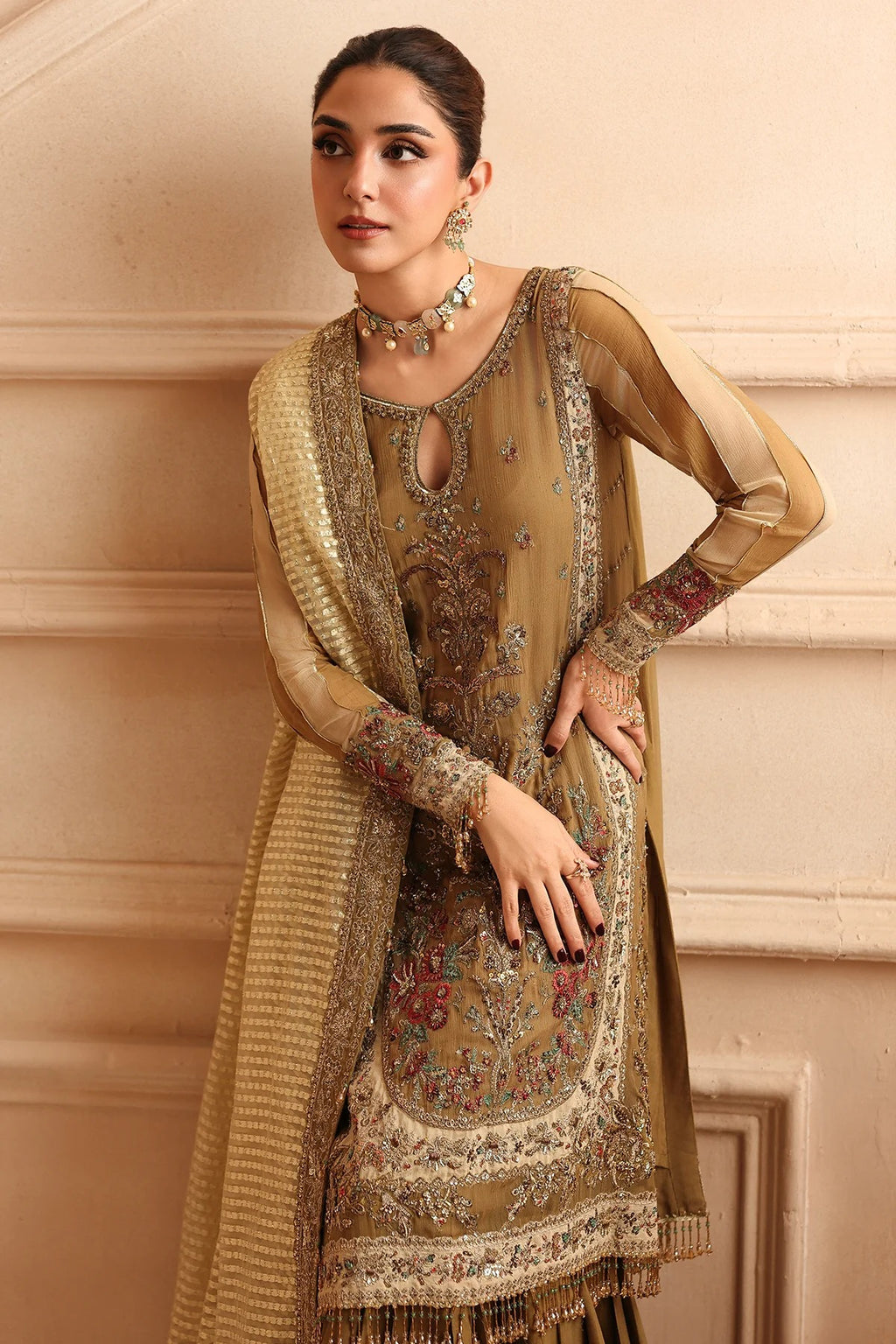 A woman models a traditional South Asian salwar kameez suit in a warm beige and gold color, featuring intricate floral embroidery and a matching textured dupatta, set against a beige stone wall background.