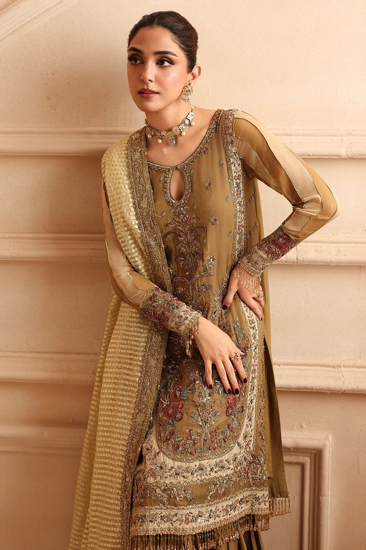 A woman models a traditional South Asian salwar kameez suit in a warm beige and gold color, featuring intricate floral embroidery and a matching textured dupatta, set against a beige stone wall background.
