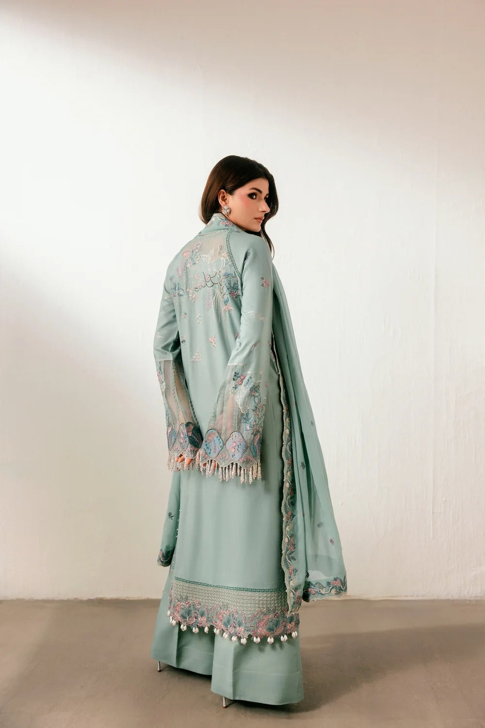 A woman wearing a mint green traditional outfit with intricate embroidery and a matching dupatta, photographed from the back against a plain off-white wall.