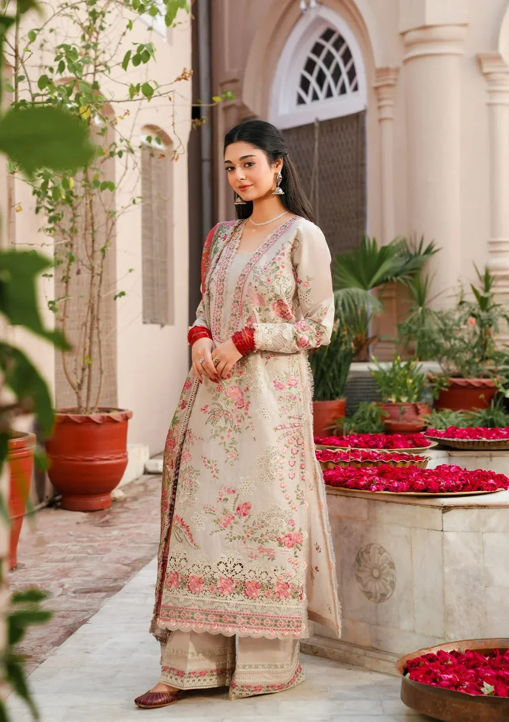 Woman in a floral traditional outfit standing in an outdoor setting with plants and a fountain.