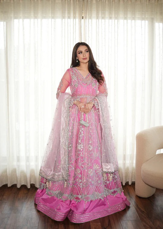 Woman wearing a pink traditional outfit with intricate designs in a room with white curtains.