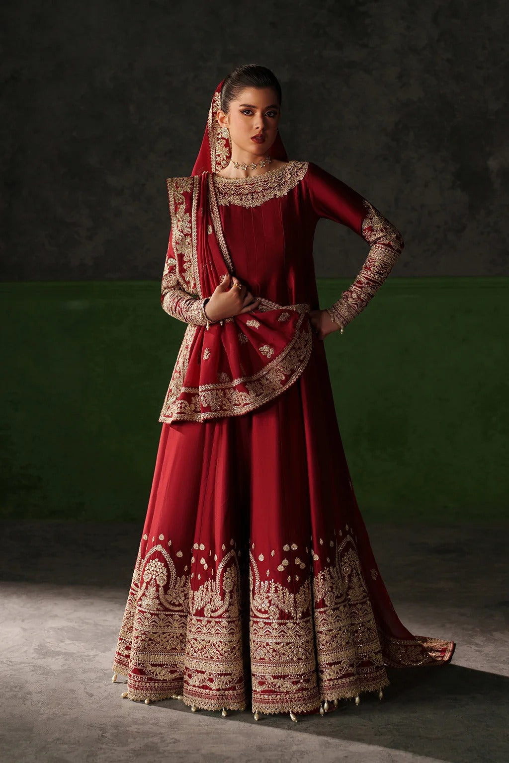 A woman models a traditional red and gold embroidered Indian outfit, featuring a long red skirt with intricate gold patterns, a matching red top with gold embroidery, and a red dupatta, set against a dark gray and green studio background.