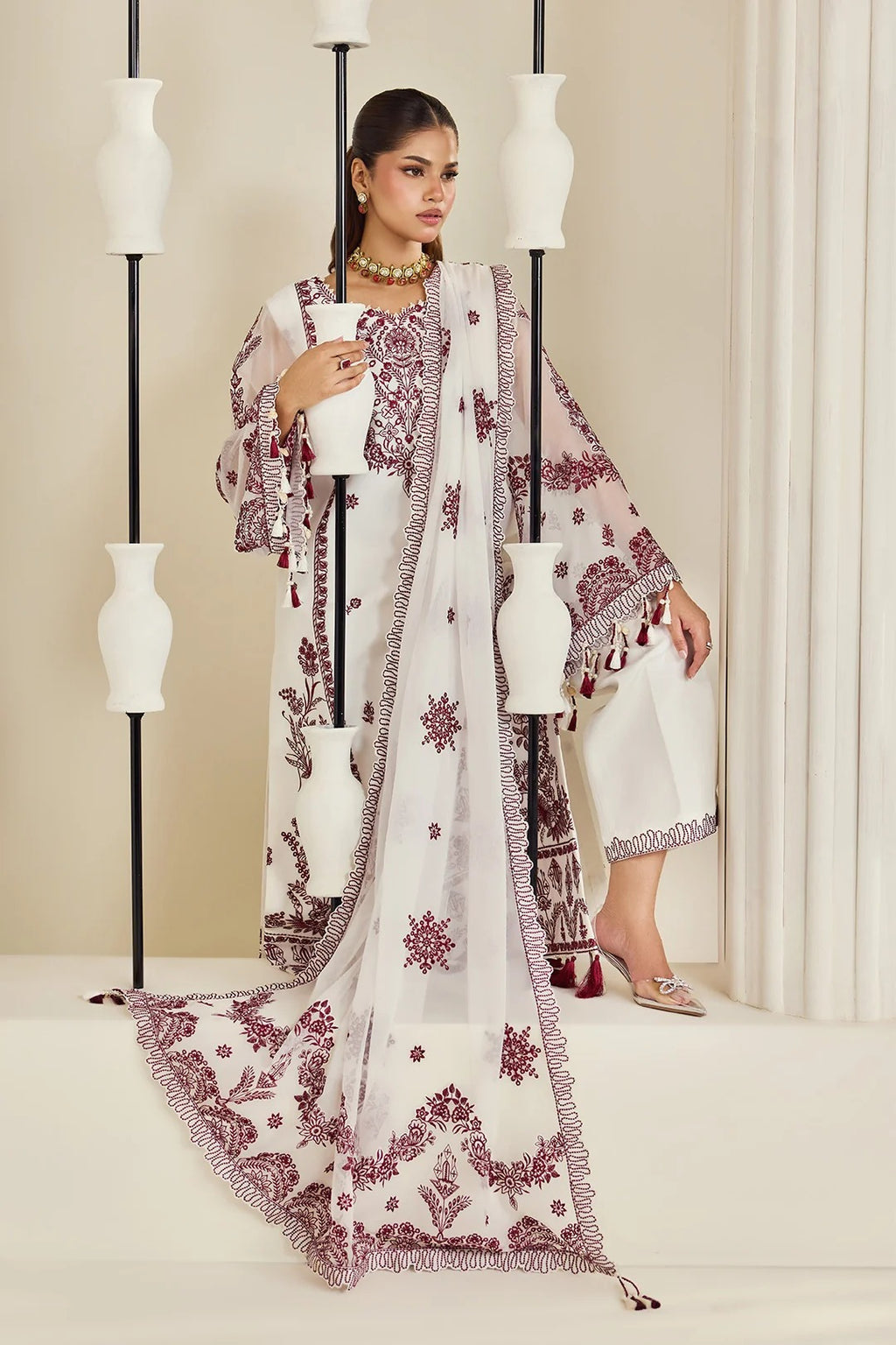 A woman models a traditional white ethnic outfit with maroon floral embroidery and border detailing, styled with a matching dupatta, against a warm beige background with decorative white vases.