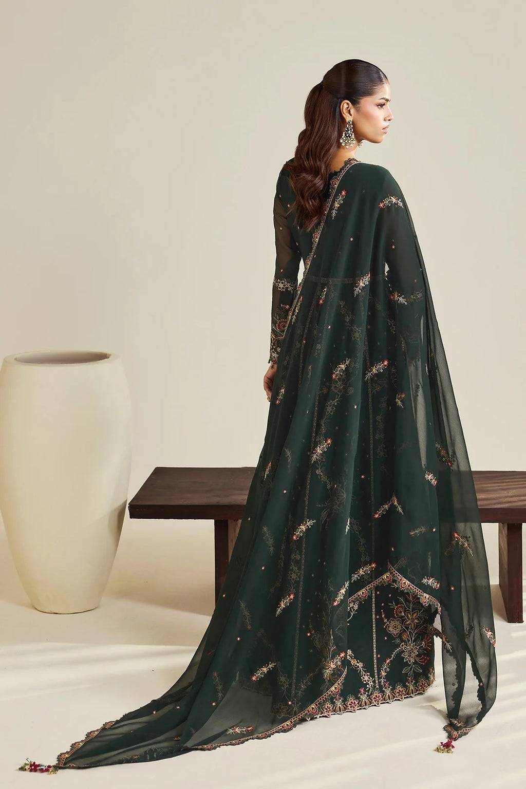 A woman modeling a dark forest green traditional outfit with intricate gold and brown floral embroidery, shown from a side-back angle in a studio setting with a beige wall, wooden bench, and white vase.
