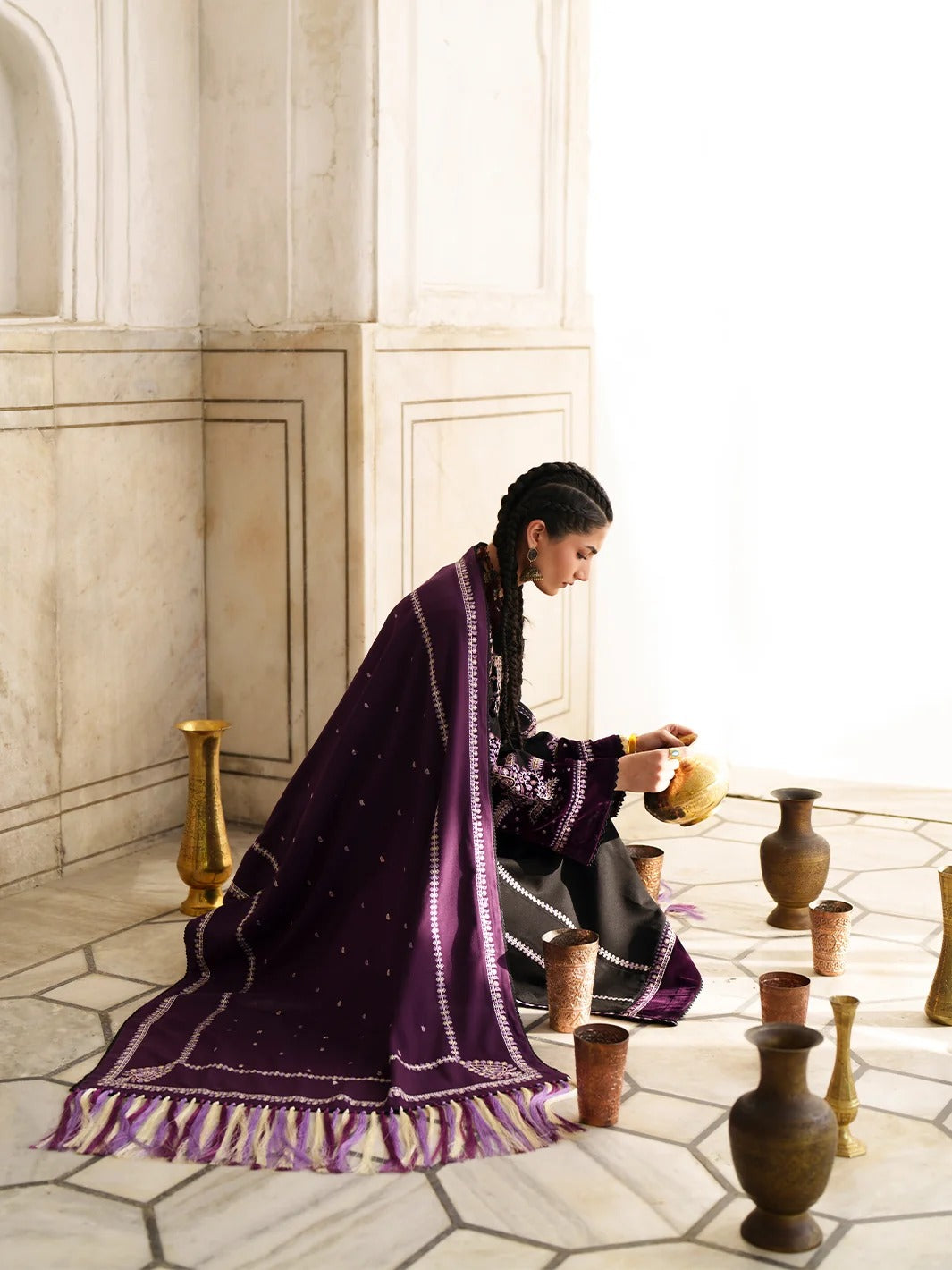 A woman in a purple embroidered shawl and black dress kneels on a stone floor, pouring from a small golden vessel among rustic clay pots and cups in a bright, classical interior.
