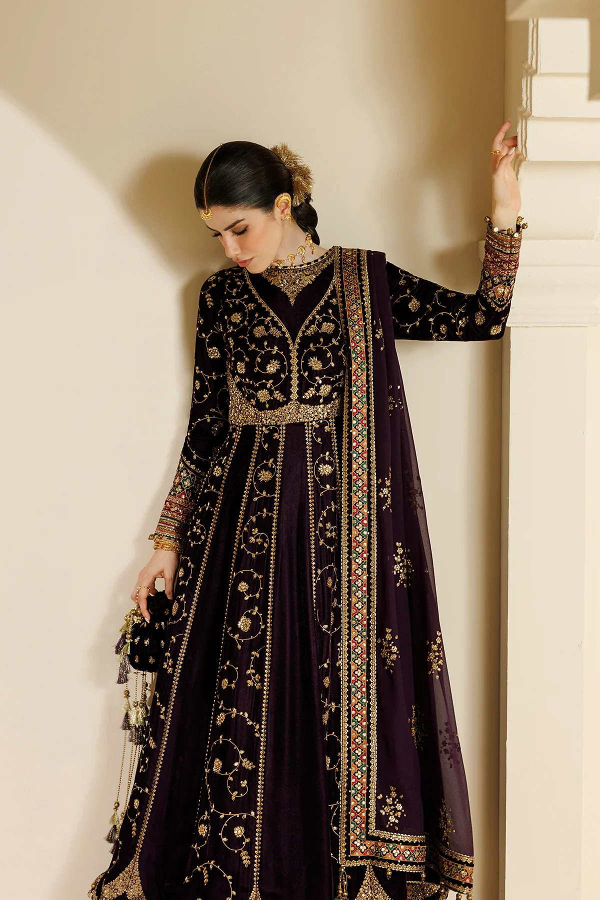 A woman models a traditional Indian outfit consisting of a deep purple velvet anarkali-style dress with extensive gold and silver embroidery, a matching embroidered dupatta, and a small black embroidered clutch bag, set against a warm beige wall.