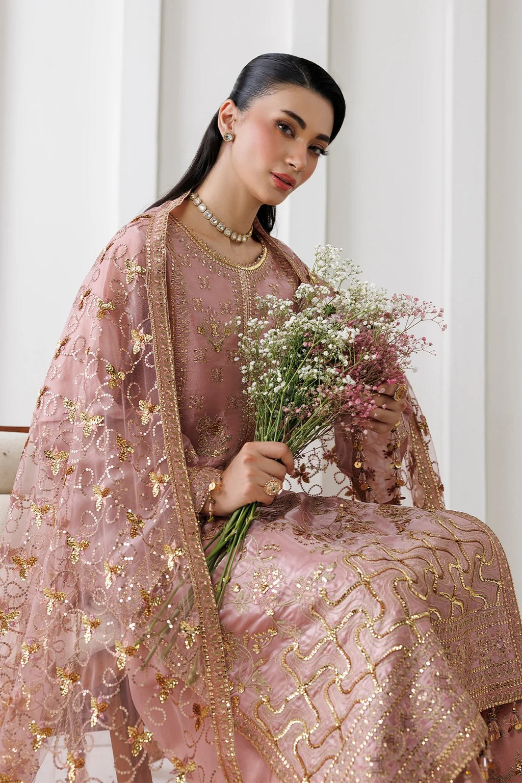 A woman wearing a pink traditional embroidered outfit with a matching sheer dupatta and gold embellishments, holding a bouquet of baby's breath flowers against a soft white background.