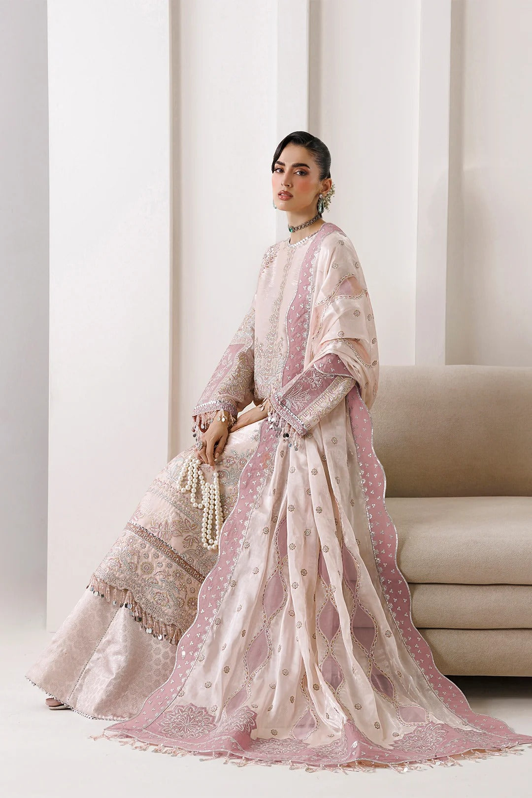A woman models a traditional light pink and beige embroidered ethnic outfit with a matching sheer dupatta in a minimalist indoor setting with a beige sofa.