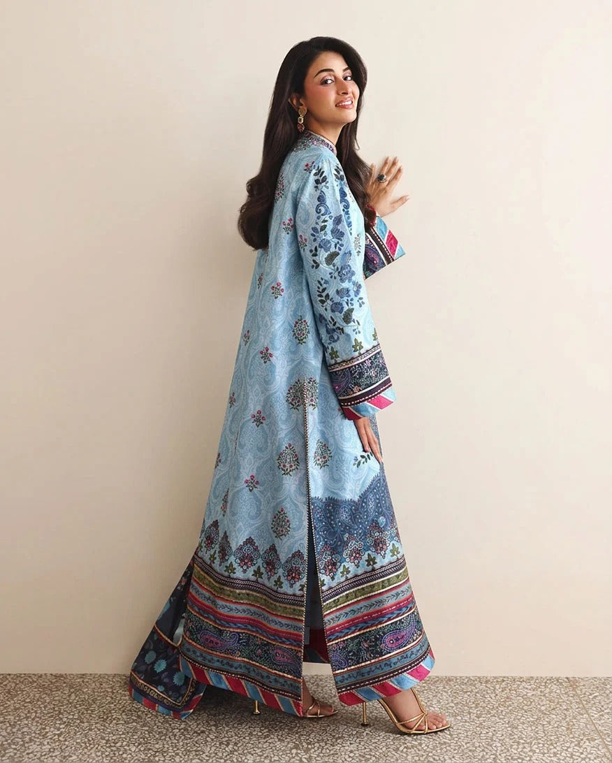 A woman modeling a light blue printed traditional long coat with colorful patterned borders, standing against a plain beige wall.