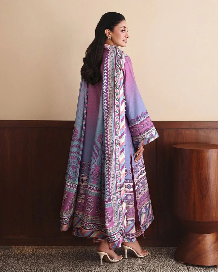 A woman wearing a long, flowing printed kaftan dress with wide sleeves, shown from the back in a studio setting with a beige wall, dark wood paneling, and a wooden side table.